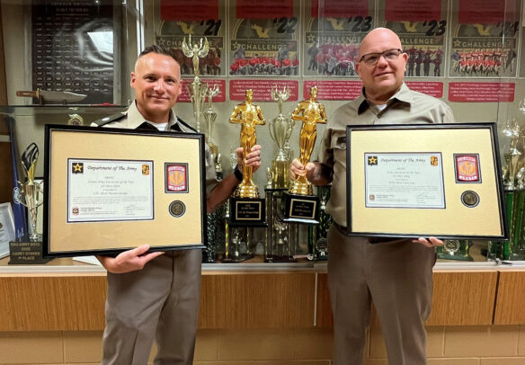 Tom Krupp and Larry Lee pose in front of an awards case holding large framed certificates and statues.
