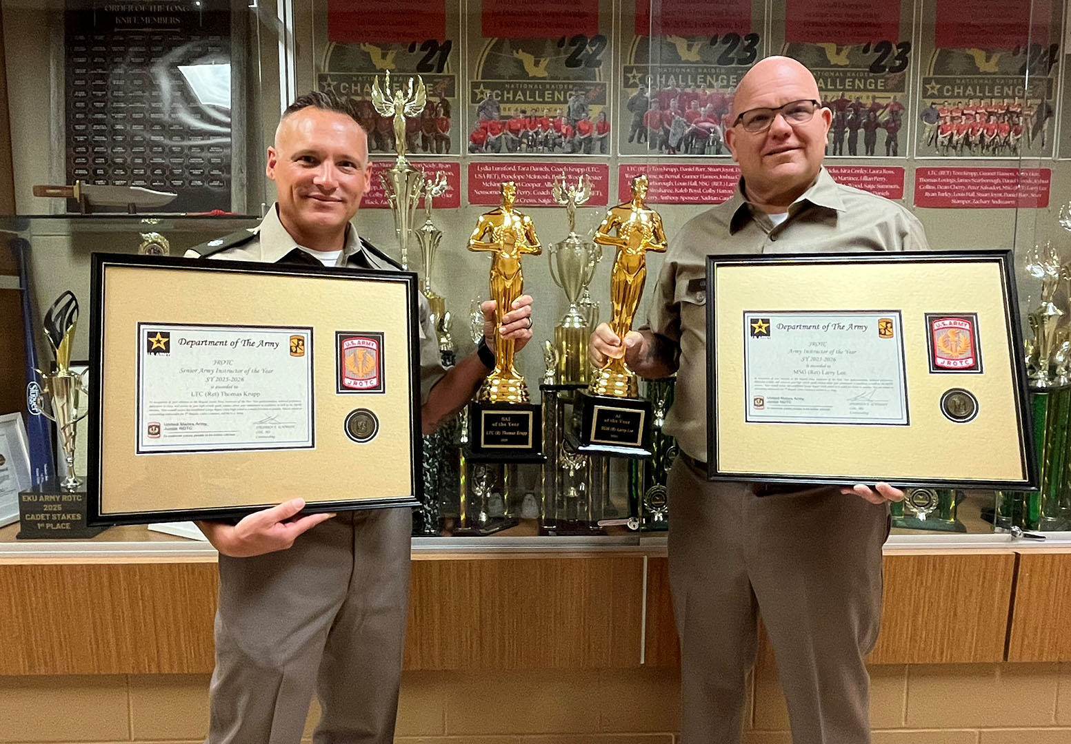 Tom Krupp and Larry Lee pose in front of an awards case holding large framed certificates and statues.