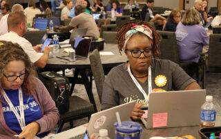 Two women sit at computers working, surrounded by tables full of teachers either working on their computers or talking.