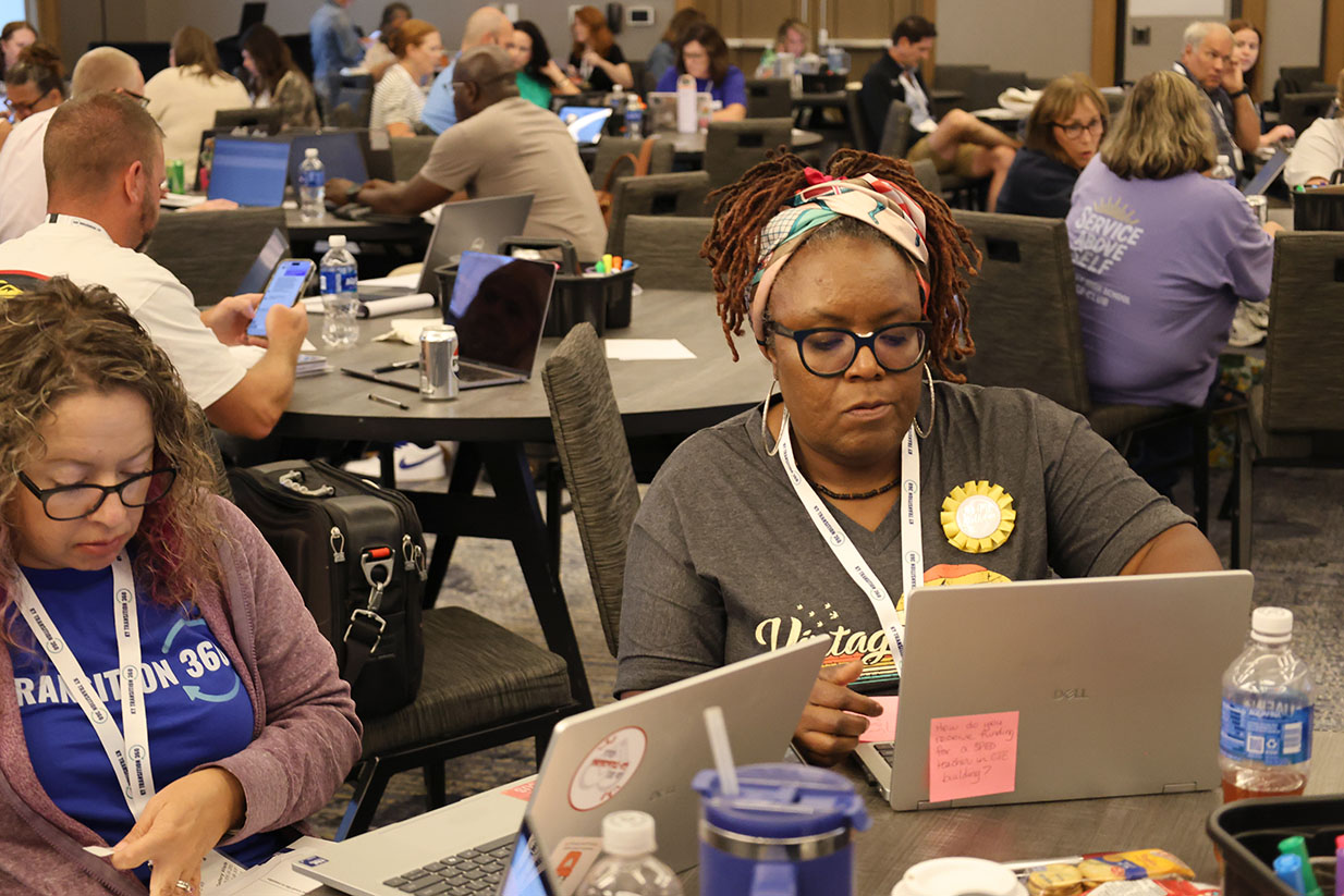 Two women sit at computers working, surrounded by tables full of teachers either working on their computers or talking.