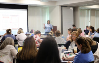 A group of teachers sit at tables, taking notes while listening to a speaker.