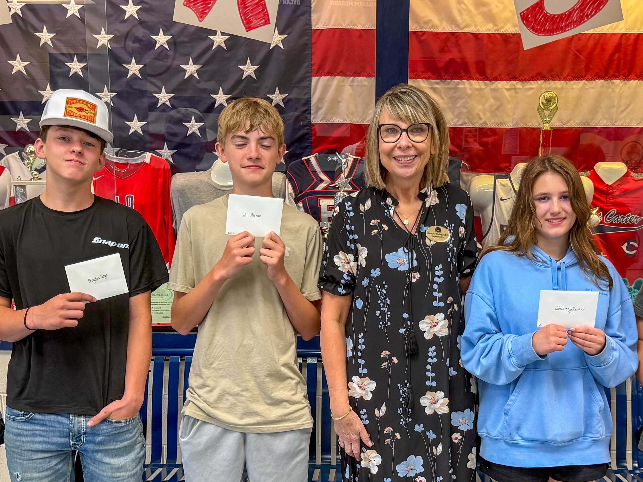 Jennifer Fraley poses for a photo in front of an American flag with three students who are holding up postcards