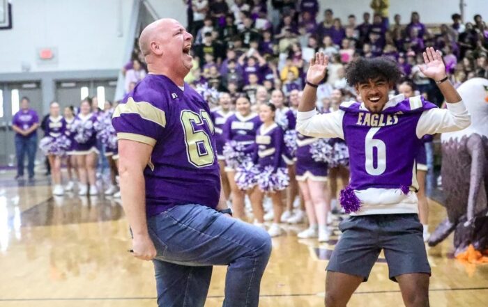 A man in a football jersey shouts while at a pep rally with other students in a gymnasium, including a student wearing a football jersey off to the side of the man