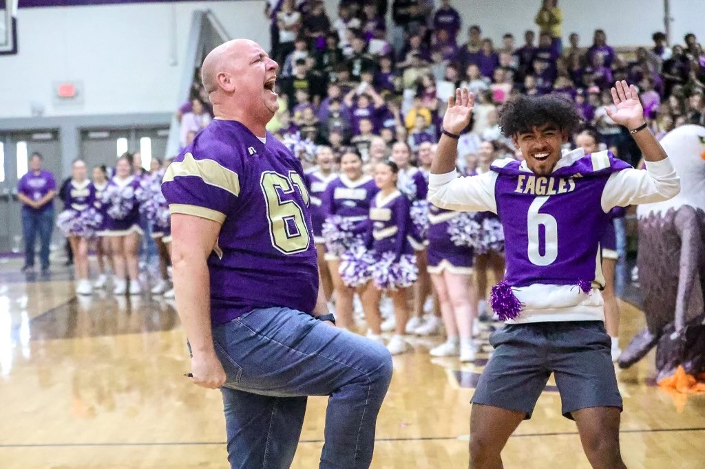 A man in a football jersey shouts while at a pep rally with other students in a gymnasium, including a student wearing a football jersey off to the side of the man