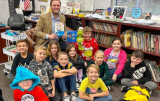 A man sits and holds a book while posing for a photo in a classroom with several other kids sitting and smiling