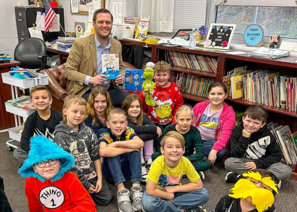 A man sits and holds a book while posing for a photo in a classroom with several other kids sitting and smiling