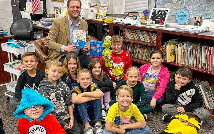 A man sits and holds a book while posing for a photo in a classroom with several other kids sitting and smiling