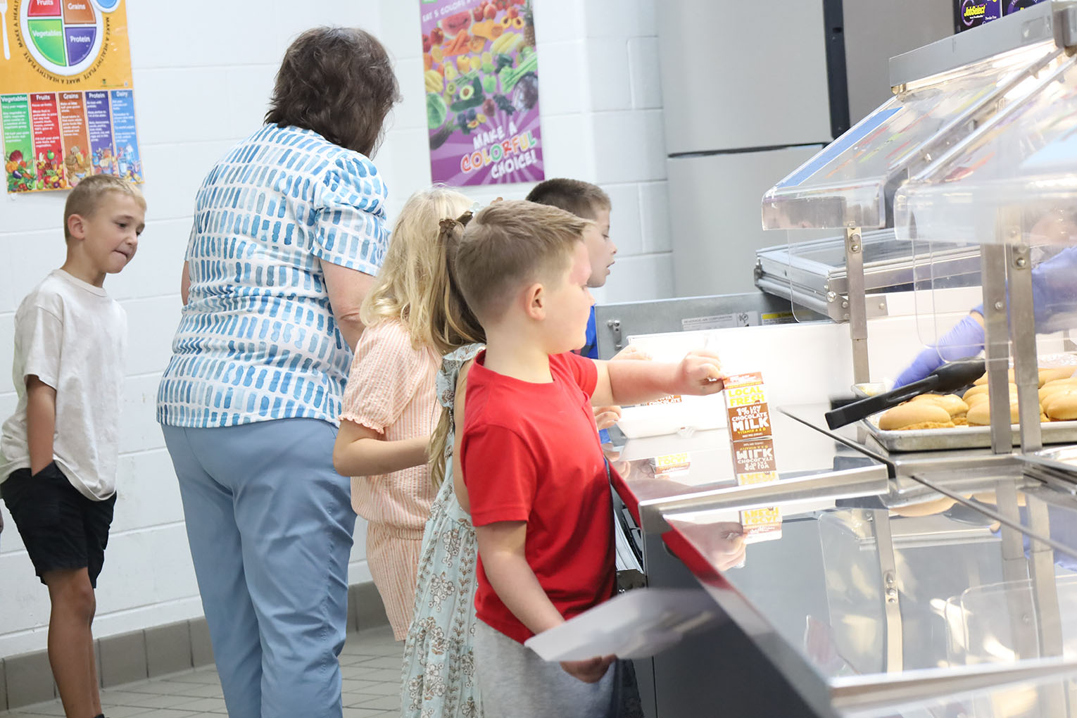 A group of young students stand in a cafeteria line waiting to pick up their lunches.