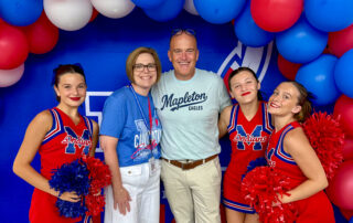 A group of people, including three girls dressed as cheerleaders around a man and a woman, smile and pose for a photo together under a red, white and blue balloon archway