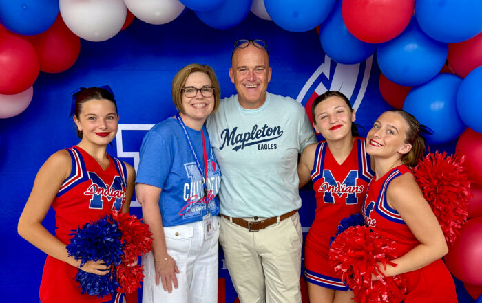 A group of people, including three girls dressed as cheerleaders around a man and a woman, smile and pose for a photo together under a red, white and blue balloon archway