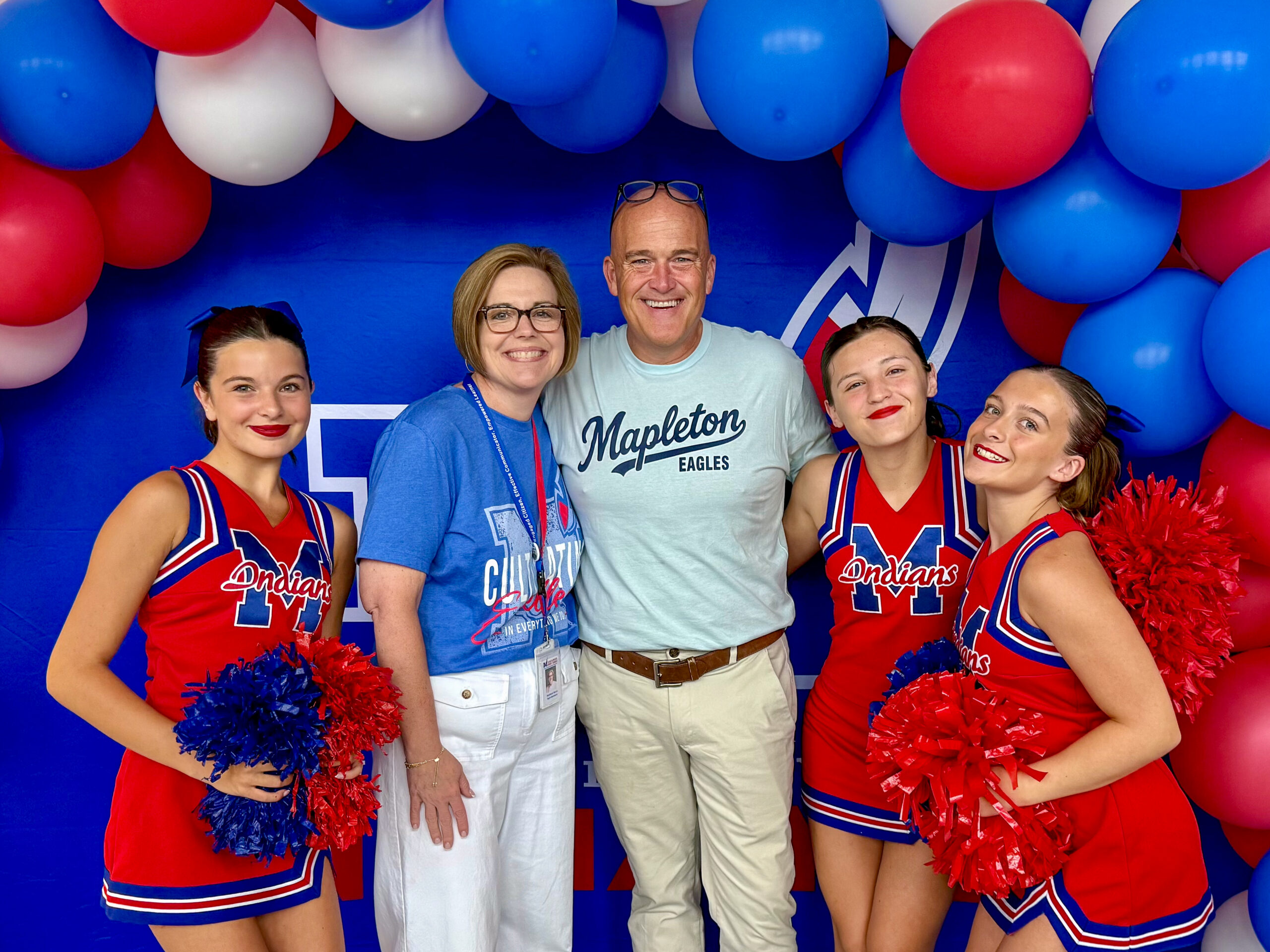A group of people, including three girls dressed as cheerleaders around a man and a woman, smile and pose for a photo together under a red, white and blue balloon archway