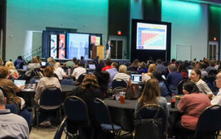 A large group of educators sit at tables in a ballroom listening to a speaker on stage in front of them.