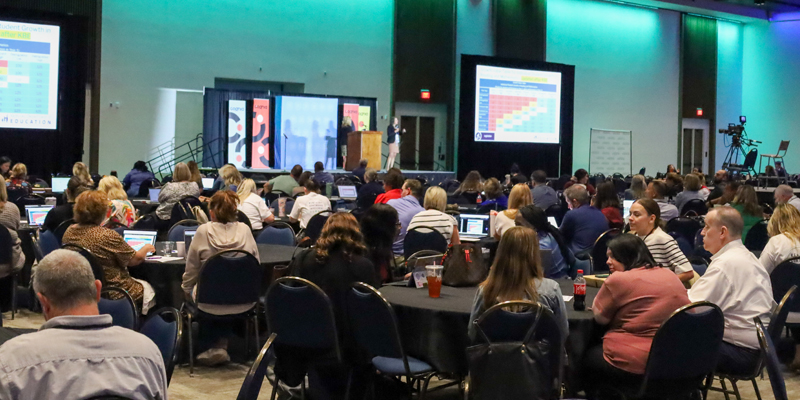 A large group of educators sit at tables in a ballroom listening to a speaker on stage in front of them.