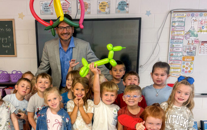 Sean Bailey poses with a group of students in a classroom with a balloon animal on his head