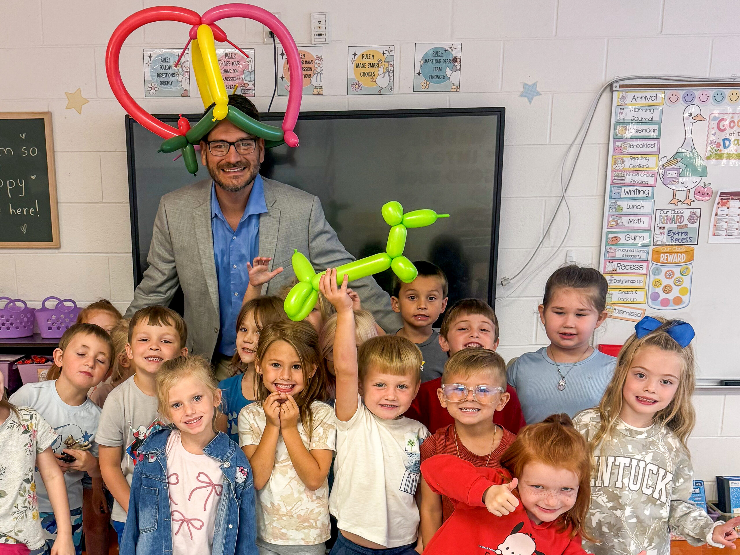 Sean Bailey poses with a group of students in a classroom with a balloon animal on his head