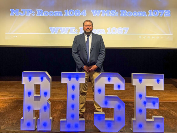 A man in a suit stands on a stage behind letters that say RISE