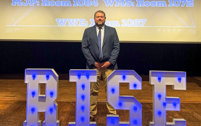 A man in a suit stands on a stage behind letters that say RISE