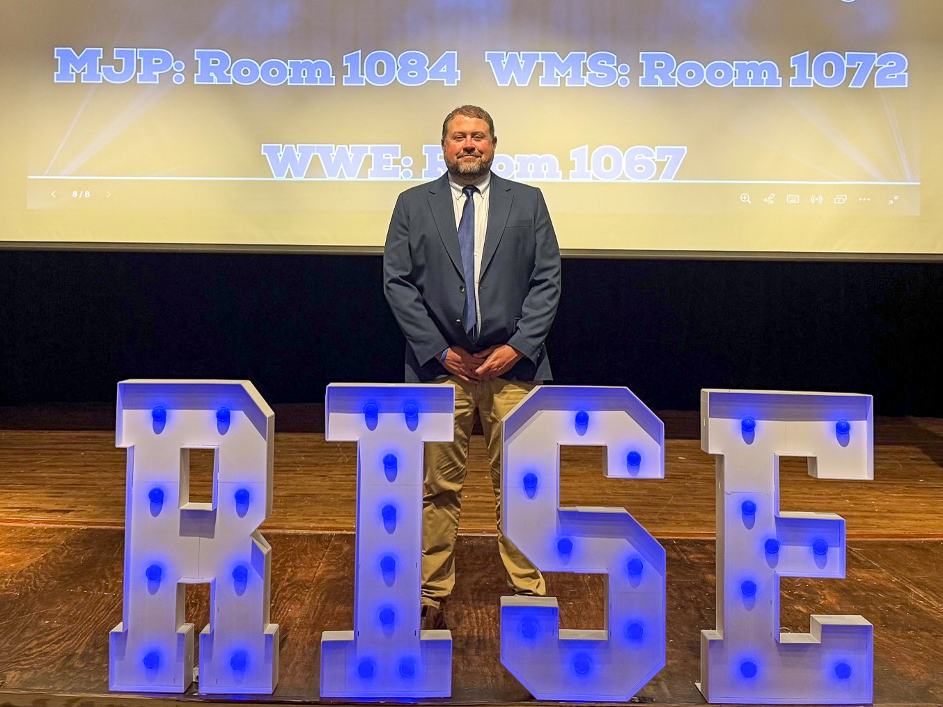 A man in a suit stands on a stage behind letters that say RISE
