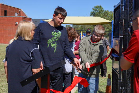 A group of kids cut a giant red ribbon in front of a metal gate