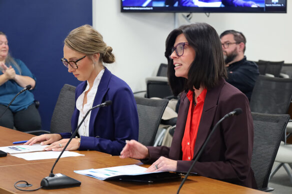 A woman is sitting at a table and talking next to another sitting woman who is looking down at pieces of paper