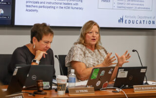 A woman sits at a table with laptops in front of her as she speaks and holds her hands out demonstratively while another woman sits next to her and looks at a screen