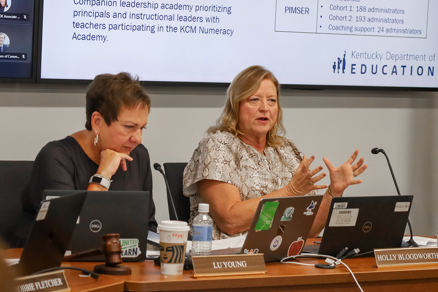 A woman sits at a table with laptops in front of her as she speaks and holds her hands out demonstratively while another woman sits next to her and looks at a screen