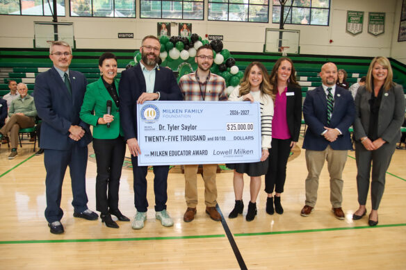 A group of people stand smiling in a gym, holding a large check made out for $25,000.